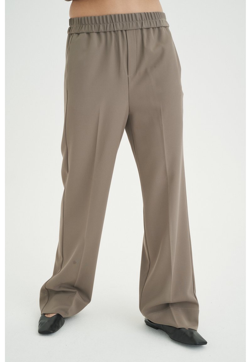 Pantalon ample taupe à taille élastique porté avec des chaussures plates noires à bouts pointus, porté par une personne debout sur fond blanc.