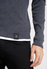Grauer Strickpullover mit runder Saumkante, versehen mit einem aufgenähten Etikett am Saum. Kombiniert mit schwarzen Jeans, zeigt eine Hand, die auf der Taille ruht.