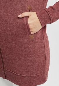 Felpa bordò con una consistenza morbida, dotata di chiusura con zip, due tasche laterali con dettagli in pelle marrone a forma di triangolo e polsini a costine.