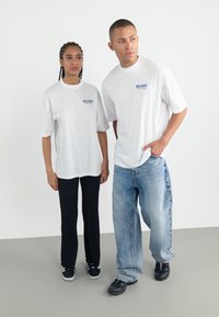 Hvide t-shirts med "DELIGHT" i blå, sorte ribbede bukser og løse lyseblå jeans. Sorte sko med farvede accenter. Minimalistisk design.