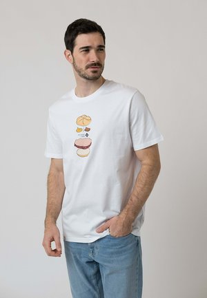 Mann trägt weißes T-Shirt mit Grafik von Sandwich-Zutaten, steht mit einer Hand in der Jeans-Tasche und schaut seitlich vor hellem Hintergrund.