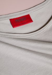 Lichtgrijs T-shirt met een ronde halslijn en zichtbare stiksels. Heeft een rood stoffen label met "HUGO" in grijze tekst. Zachte textuur.