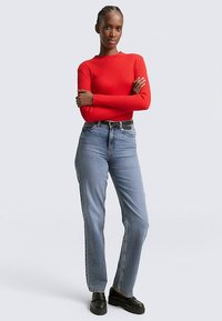 Rotes, geripptes Langarmshirt, hochtaillierte hellblaue Jeans, schwarzer Gürtel und schwarze Schuhe, mit verschränkten Armen vor einem einfarbigen Hintergrund stehend.