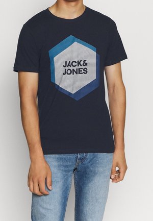 Hombre vistiendo una camiseta negra con un logo hexagonal azul y el texto "JACK & JONES", combinada con jeans de un azul claro.