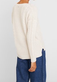 Beżowy sweter dzianinowy z teksturowymi wzorami, z detalami w warkocz na rękawach i bocznymi rozcięciami. Stworzono go w połączeniu z niebieskimi spodniami dżinsowymi.