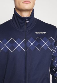 Hombre que lleva una chaqueta Adidas de color azul marino con un patrón de diamantes en azul y blanco y un cuello alto con cremallera, cortada por debajo de la boca.