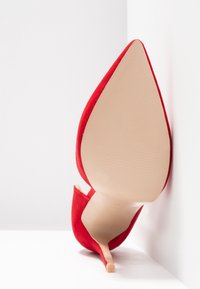 Chaussure à talon haut en daim rouge avec un bout pointu et un talon stiletto mince. La semelle est beige lisse avec des motifs texturés pour l'adhérence.