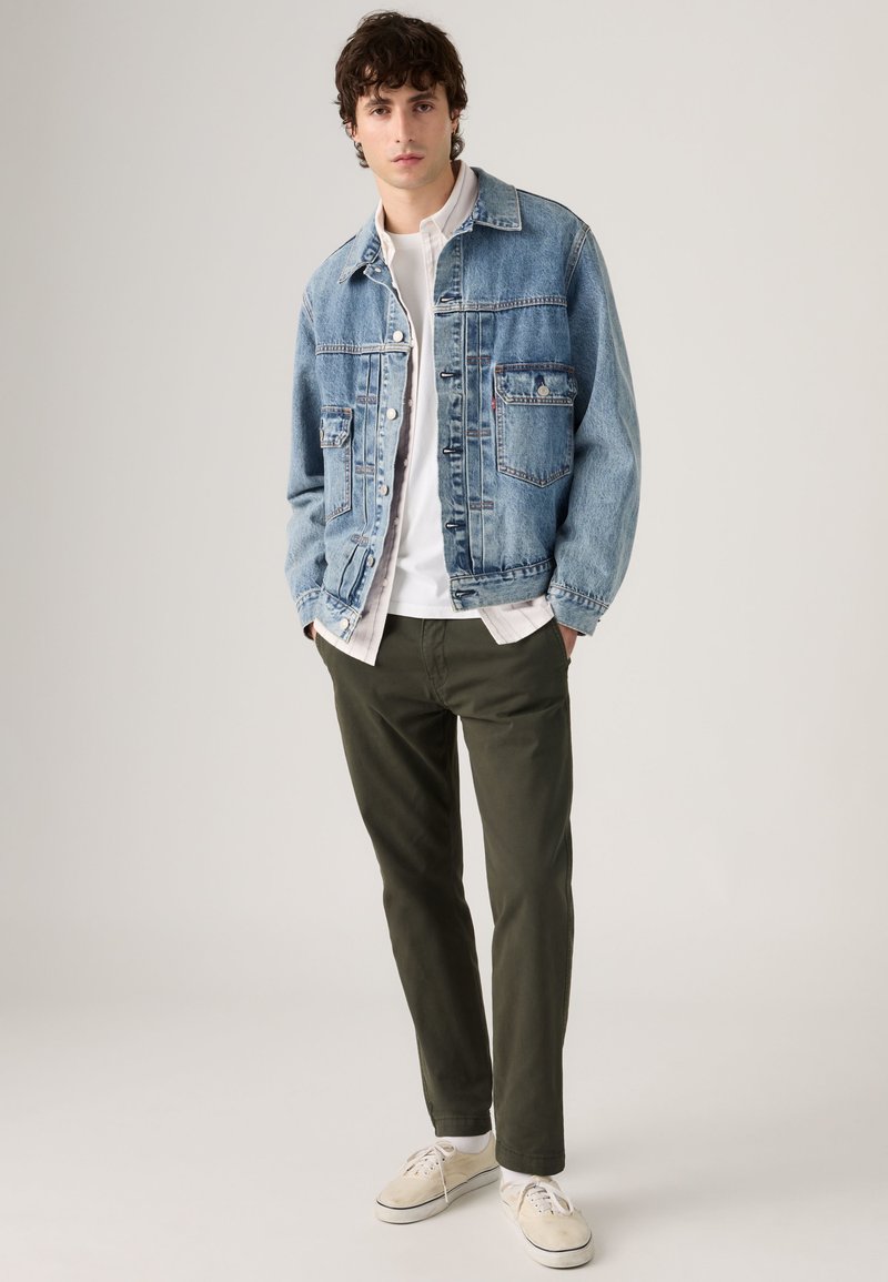 Jeune homme debout, les mains dans les poches, portant une veste en jean bleu clair, une chemise blanche, un pantalon vert olive et des baskets beige.