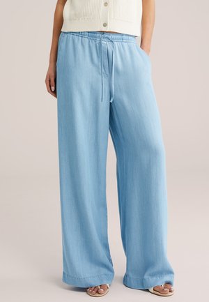 Kobieta ubrana w jasnoniebieskie szerokie spodnie z elastycznym pasem i sznurkiem, zestawione z białymi sandałami na otwarte palce oraz kremowym dzianinowym topem bez rękawów.