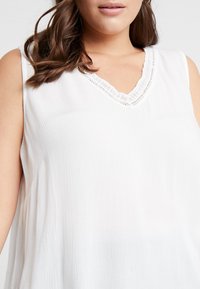 Top bianco senza maniche con scollatura a V, caratterizzato da un tessuto testurizzato e un delicato bordino in pizzo lungo il collo. Design leggero e informale.