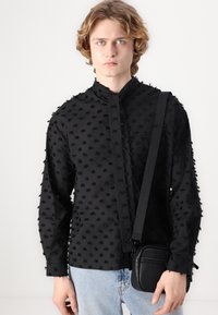 Black textured shirt with 3D dot pattern, high collar, button closure, and long sleeves. Worn with a small black crossbody bag.