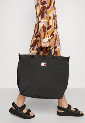 Black nylon tote bag with a rectangular shape, dual top handles, and a logo patch. Worn with a patterned dress and black sandals.