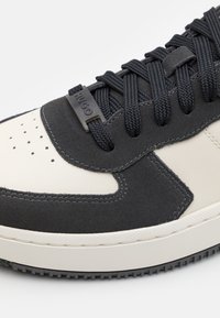 Gros plan sur l'avant d'une basket noire et blanche avec des lacets noirs et une étiquette de lacet marquée "HUGO" sur des panneaux en suède texturé et en cuir.