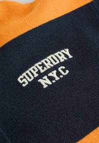 Gebreide trui met marineblauwe en oranje strepen. Kenmerkt zich door witte geborduurde tekst "SUPERDRY N.Y.C." op een donkere achtergrond. Zachte textuur.