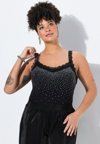 Top negro sin mangas con detalles de encaje y brillantes tachuelas plateadas, combinado con pantalones negros plisados y brillantes. La mano del modelo descansa en la cadera.