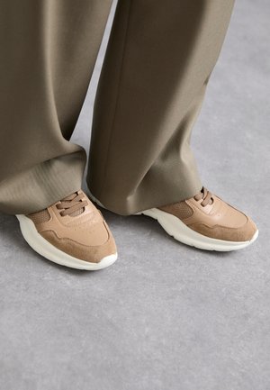 Sapatilhas desportivas castanhas com uma mistura de pele suave e materiais em malha, com uma sola de borracha branca e discretos detalhes de perfuração.