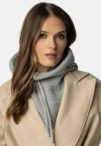 Cappotto di lana beige sopra un hoodie grigio chiaro, a trama. L'hoodie presenta un cordino e un grande cappuccio, offrendo un look a strati.