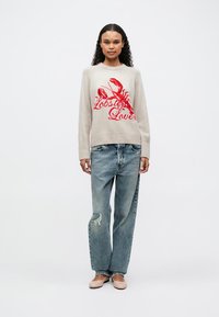 Pull en tricot beige avec un motif de homard rouge et le texte "Amateur de homard". Porté avec un jean bleu déchiré et des chaussures de couleur claire.