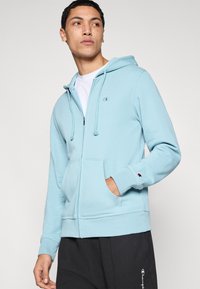 Φωτεινό γαλάζιο φούτερ με φερμουάρ, φτιαγμένο από απαλό υλικό, με τσέπη μπροστά, κουκούλα με κορδόνια και μικρό λογότυπο στο στήθος.