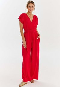 Czerwony kombinezon z krótkimi rękawami, głębokim dekoltem w kształcie litery V, paskiem w talii i szerokimi nogawkami. Gładki materiał o miękkiej teksturze i bez widocznych wzorów.