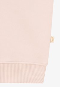 Sweatshirt rose clair en tissu doux, avec des poignets côtelés et une petite étiquette de marque beige. Texture lisse avec un design simple et minimaliste.