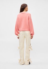 Ροζ άνετος πουλόβερ με χαλαρή εφαρμογή, συνδυασμένος με κρεμ παντελόνια που διαθέτουν φιόγκο στα μανίκια. Απλό φόντο.