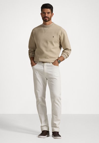 Beige genser med rund hals, hvite jeans og brune sneakers. Har ribbestrikkede mansjetter og en liten logo på brystet. Ledig passform og lett stoff.