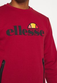 Sweat-shirt rouge avec un grand logo "ellesse" noir et un accent en demi-cercle jaune et rose. Comprend des zips sur les côtés et un col rond.