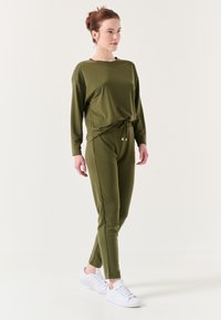 Olijfgroene loungewear set met een ontspannen fit long-sleeve top en broek met koord, voorzien van een zachte stof en subtiele accenten aan de zijkant. Gecombineerd met witte sneakers.