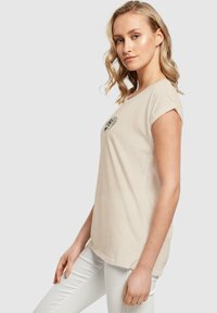 Hellbeige Baumwoll-T-Shirt mit Rundhalsausschnitt und kurzen hochgeschlagenen Ärmeln. Auf der linken Brustseite befindet sich ein schwarzes grafisches Emblem.