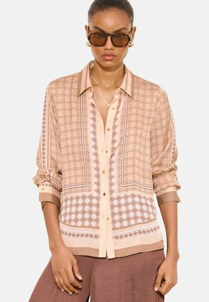 Femeie care poartă o bluză bej cu nasturi și model, cu mâneci suflecate, ochelari de soare maro, cercei aurii de tip cerc, colier și pantaloni maro.