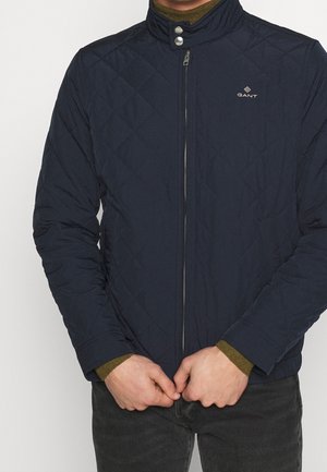 Mand iført en navyblå quiltet jakke med lynlås og trykknap-krave, hænderne foldet foran, olivengrønne sweaterærmer synlige.