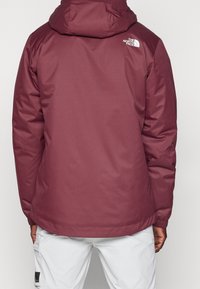 Bordowa, ocieplana kurtka z kapturem, wykonana z teksturowanej tkaniny i z białym logo na górnej części pleców, zaprojektowana w celu ochrony przed zimnem.