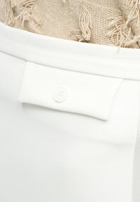 Κοντινό πλάνο λευκών παντελονιών με πλαϊνή τσέπη με κουμπιά, συνδυασμένα με μπεζ πλεκτό τοπ με υφή.