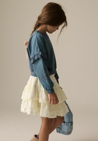 Blauwe sweatshirt met kantaccenten en bloemenborduursel, gecombineerd met een gelaagde witte ruchesrok. Denim tas in de rechterhand.