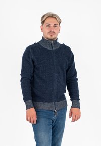 Maglione zip-up blu navy con colletto alto, accenti a coste grigio chiaro sui polsini e sull'orlo, realizzato in tessuto testurizzato. Indossato con jeans blu.