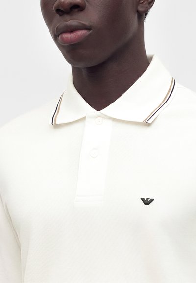 Homme portant un polo blanc avec une patte à deux boutons et un petit logo noir brodé sur la poitrine.