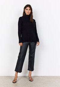 Schwarzer gerippter Rollkragenpullover mit langen Ärmeln, kombiniert mit schwarzen weiten Hosen aus Kunstleder und schwarzen Riemchensandalen. Schlichte Accessoires.