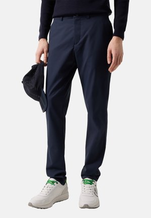 Mand iført marineblå bukser og sort sweater, der holder en sort kasket i venstre hånd, med hvide sneakers med grønne snørebånd.