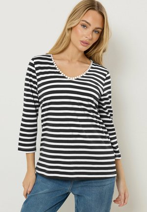 Blonde Frau, die ein schwarz-weiß gestreiftes Langarmshirt mit Wellenkante am Halsausschnitt und blaue Jeans trägt, vor weißem Hintergrund stehend.