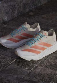 Pair of beige running shoes with orange stripes and light blue laces on a stone pavement surface.