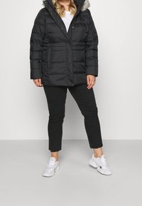 Persona con chaqueta de plumas negra y capucha con piel sintética, pantalones cortos negros, zapatillas blancas, de pie contra un fondo claro y uniforme.