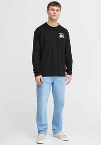 Black long-sleeve t-shirt with a graphic print on the left chest, paired with light blue jeans and beige sneakers. Smooth fabric, casual fit.