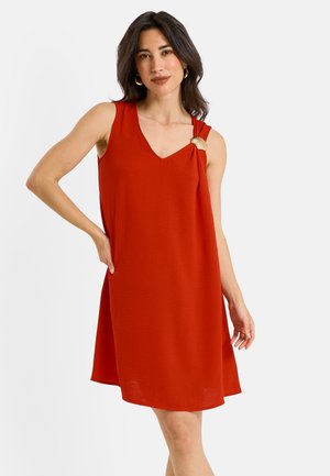 Femme aux cheveux foncés portant une robe rouge sans manches avec un fermoir doré en forme de coquillage sur l'épaule, debout avec la main sur la hanche sur fond blanc.