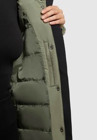 Pufota jaka ar melnu ārējo virsmu un olīvu zaļu iekšējo oderi. Redzami pogu sprādzes un sānu kabata.