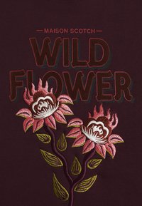 El tejido burdeos presenta el texto 'WILD FLOWER' en rojo y rosa, con flores rosas bordadas y hojas verdes, mostrando un detallado trabajo de costura.