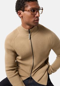 Beiger Reißverschluss-Pullover mit gerippten Akzenten an Schultern und Bündchen, mit hohem Kragen und weicher Textur. Getragen über dunklen Jeans.