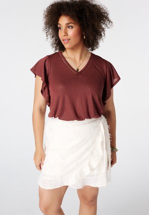 Femme aux cheveux bouclés portant un haut à manches volantes rouge foncé et une jupe portefeuille blanche en tissu texturé, regardant vers sa gauche.