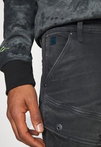 Högra handen nära fickan på svarta jeans med en logopatch, kombinerat med en svart och grå långärmad tröja med manschett.