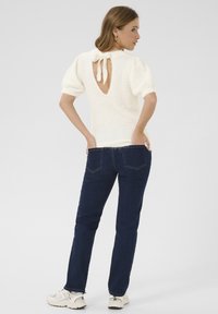 Pull en tricot blanc avec des manches courtes bouffantes et un détail à nouer à l'arrière, assorti à un jean slim fit bleu foncé et des baskets blanches.